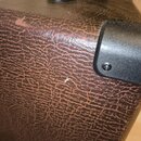 Detail hnědého povrchu komba potaženého texturou imitující kůži. V pravé části je vidět černý plastový roh připevněný šroubem. Povrch vykazuje známky…