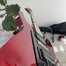 Kytara s červeným lakovaným tělem a hnedým hmatníkem. Nástroj má dva černé humbuckery a černý pickguard. Na fotce nejsou žádné viditelné značky ani n…