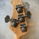 Zadní strana hlavy basové kytary z přírodního dřeva. Na hlavě jsou čtyři chromované ladičky, z nichž každá nese nápis MUSIC MAN. Povrch dřeva i kovov…