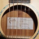 Detail zvukového otvoru akustické kytary s perleťovým lemováním. Uvnitř je nalepený štítek s logem Faith a nápisem Faith Guitars. Štítek obsahuje tex…