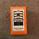 Krabice oranžové barvy s bílým štítkem, na kterém je zobrazen kytarový pedál. Na štítku jsou nápisy SIGNAL PAD, PASSIVE ATTENUATOR a značka electro-h…