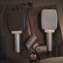 Dvě mikrofony s mřížkou a jeden držák v černých látkových pytlích. Levý mikrofon je černý s nápisem SENNHEISER e606 a MADE IN GERMANY, v dolní části…