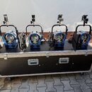 Čtyři modré reflektory s černými klapy a úchyty jsou umístěny na černé transportní bedně s hliníkovými hranami a koly. Na každém reflektoru je vidite…