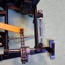 Detailní pohled na mechanismus černého pedálu s žlutým praporkem. Součástí je kovový řetěz, pružina a nastavovací šroub. Na komponentech nejsou žádné…