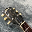 Detail hlavy černé kytary s nápisy Gibson, MODEL Les Paul a Deluxe. Nástroj má bílé ladičky a hnědý hmatník s kovovými pražci. Povrch hlavy vykazuje…