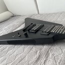 Černá elektrická kytara s matným povrchem a hranatým, asymetrickým tvarem těla. Nástroj má dva humbuckery, jeden ovládací knoflík a přepínač. Na těle…