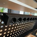 Černá kovová lišta s řadou deseti kulatých otvorů pro zapojení kabelů. Lišta je připevněna šrouby k perforovanému černému kovovému panelu. Na kompone…