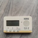 Bílý chromatický ladič značky KORG, model CA-2. Zařízení má digitální displej a mřížku reproduktoru. Na spodní straně jsou tlačítka s nápisy CALIB, A…