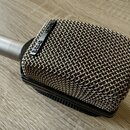 Kovový mikrofon s obdélníkovou mřížkou a stříbrným hrdlem. Na černé liště u mřížky je bílý nápis AKG D12E. Povrch mřížky vykazuje drobné známky opotř…