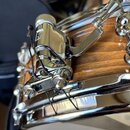 Detailní záběr na chromovaný mechanismus napnutí plachet u bubnu s dřevěným korpusem v hnědé barvě. Na kovovém dílu je vyryt symbol trojúhelníku. Pov…