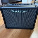Černý kytarový kombo zesilovač s černou textilní mřížkou. Uprostřed mřížky je bílý nápis Blackstar. Na horní straně jsou vidět ovládací knoflíky a úc…