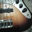 Detail elektrické basové kytary s tělem v provedení sunburst. Nástroj má světle šedý pickguard s mramorovaným vzorem a kovový panel se třemi černými…
