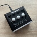 Černý kompaktní přístroj značky behringer s třemi bílými otočnými knoflíky. Na spodní straně jsou viditelné nápisy MIC/LINE GAIN 1, INST GAIN 2 a OUT…