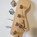 Zadní strana hlavy basové kytary z přírodního světlého dřeva s chromovanými ladičkami. Na dřevě je černý nápis Fender® JAZZ BASS®. Povrch dřeva a kov…