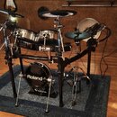 Elektronická bicí souprava značky Roland umístěná na modrém koberci. Na basovém bubnu je bílý nápis Roland a níže označení V-Drums. Souprava obsahuje…