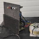 Fotografie zobrazuje elektronické zařízení v rozloženém stavu, pravděpodobně zesilovač nebo napájecí zdroj. Sestava obsahuje černý kovový panel s ovl…