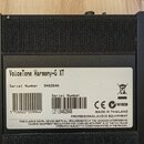 Fotografie zachycuje zadní stranu černého zařízení s texturovaným povrchem. Na bílé nálepce je uveden název VoiceTone Harmony-G XT a sériové číslo 94…