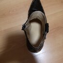 Na fotografii je černá botka s špičatou zástupní částí, položená na dřevěné podlaze. Vnitřek botky je světle béžový a v oblasti paty je viditelné opo…