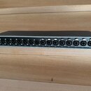 Šedý rackový audio přístroj s černým horním krytem. Na zadním panelu jsou umístěny konektory XLR a dva konektory Ethernet s nápisy SEND a RETURN. V l…