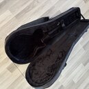 Otevřený černý pouzdro na kytaru s vnitřním polstrováním z černého plyšového materiálu. Pouzdro je rozloženo na světlé laminátové podlaze. Na vnitřní…
