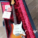 Elektrická kytara s tónem sunburst a bílým pickguardem v otevřeném kufru s červeným vnitřkem. Na hlavě kytary je logo Fender a podpis. K nástroji jso…