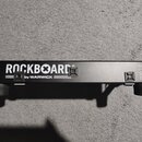 Černý kovový pedalboard s gumovými nožkami. Na horní straně je bílý nápis ROCKBOARD® by WARWICK a v pravém kraji označení DUO 2.0. Na povrchu jsou um…