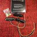 Tři černé humbuckery s viditelnými bílými póly a připojovacími vodiči v různých barvách. Vedle nich je průhledný sáček s montážním příslušením a krab…