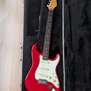 Červená elektrická kytara s bílým pickguardem a třemi snímači, umístěná v černém tvrdém kufru. Na hlavě hředu je viditelné logo Fender Stratocaster a…