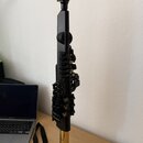 Černý dřevěný nebo plastový klarinet s kovovými zlatými klapy a zlatým zvonem. Nástroj stojí vertikálně na dřevěném stole vedle otevřeného notebooku…
