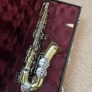 Zlatý saxofon v otevřeném černém kufru s červeným plyšovým vnitřkem. Na těle nástroje je viditelný nápis "YAMAHA". Povrch instrumentu vykazuje známky…