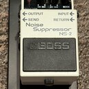 Bílý efekt pedál s černým spínacím tlačítkem a nápisem BOSS. Na přední straně je text Noise Suppressor NS-2 a označení vstupů OUTPUT, INPUT, SEND a R…