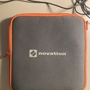 Štvrubní šedý přepravní pouzdro s oranžovým zipem. Uprostřed je bílým tiskem logo a nápis novation®. Povrch materiálu vykazuje mírné znečištění v pod…