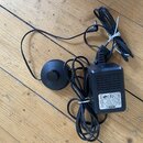 Černý síťový adaptér s připojeným kabelem a kulatým ovládacím knoflíkem na kabelu. Na štítku adaptéru je uveden nápis AC-ADAPTOR, MODEL: SV-9200A-GS,…