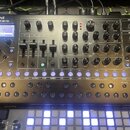 Černý syntetizátor s nápisem Roland SYNTHESIZER SH-4d. Přístroj obsahuje množství otočných knoflíků, posuvníků a řadu 16 podsvícených modrých tlačíte…
