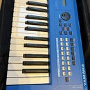 Modrý syntezátor s klaviaturaí, umístěný v černém pouzdře. Na pravé straně je svisle napsáno YAMAHA a v dolní části nápis MUSIC SYNTHESIZER mx49 spol…