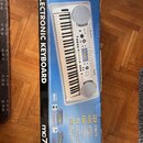 Krabice s elektronickým klavírem v modré a bílé barvě. Na boku je nápis ELECTRONIC KEYBOARD, logo midi a označení modelu mc70. V dolní části jsou uve…