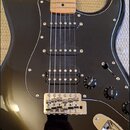 Černá elektrická kytara s tvarem typu Stratocaster, světlým javorovým hmatníkem a černým pickguardem. Nástroj má tři snímače typu single-coil, chromo…