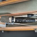 V dřevěném kufru s pěnovou výplní leží mikrofon v kovovém provedení s černým povrchem těla, který vykazuje výrazné opotřebení a odloučení barvy. Na s…