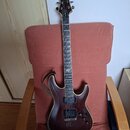 Elektrická kytara s tmavě hnědým průhledným lakem a viditnou dřevěnou strukturou. Na hlavě je bílé logo Schecter, pod ním nápis C-1. Hриф má tmavý hm…
