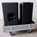 Dvě černé reproduktorové bedny umístěné v hliníkovém transportním kufru na modrých kolečkách. Na levé bedně je v oblasti úchytu bílý nápis Turbosound…
