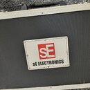 Černý transportní kufr s hliníkovým rámem a kovovými rohy. Uprostřed je připevněna bílá tabulky s červeným logem SE a nápisem SE ELECTRONICS. Povrch…