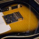 Detail elektrické kytary v barvě sunburst s bílým pickguardem, položená v kufru. Na kytaru jsou vidět dva stříbrné ovládací knofíky a kovový most s h…