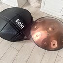 Na fotografii je měděný steelpan s několika vyhlubecemi na povrchu a černý polstrovaný přepravní vak. Na vaku je bílý nápis Sela a pod ním menší text…