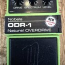 Zelený kytarový efekt s nápisy Nobels ODR-1 Natural OVERDRIVE. Na horní části jsou tři černé otočné ovladače s popisky DRIVE, SPECTRUM a LEVEL. V pra…