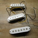 Tři bílé humbuckery s kovovými póly, položené na dřevěném povrchu. Každý snímač má připojené elektrické vodiče v různých barvách (černá, bílá, žlutá)…