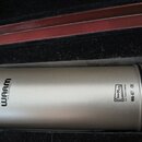 Kondenzátorový mikrofon stříbrné barvy s matným povrchem, umístěný v černé polstrované přepravce. Na těle je černý nápis WARM AUDIO a logo IneMaq s d…