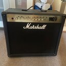 Černý kytarový kombo zesilovač s látkovou mřížkou. Na přední straně je bílé logo Marshall a na ovládacím panelu nápis MG 100W. Panel obsahuje řadu ot…