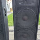 Dvě černé reproduktorové bedny s kovovou mřížkou. Na spodní části pravého reproduktoru je oranžový štítek s bílým nápisem JBL a pod ním menší nečitel…