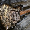 Elektrická kytara s černým lakem a výrazným zlatavě-šedým vzorem paisley na těle a pickguardu. Nástroj má hnědý hmatník, tři snímače a třy čtyři ovlá…