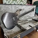 Elektrická kytara s tmavě šedým třpytivým povrchem v černém tvrdém kufru s šedým polstřením. Kytara má dva humbuckery, šest strun a černý hmatník. Na…